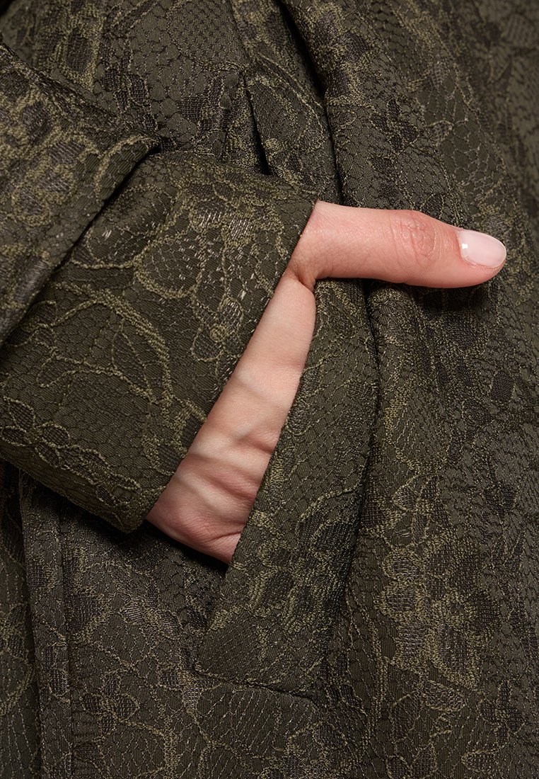 Una mano con smalto rosa pallido infilata nella tasca di una manica di tessuto verde oliva scuro con motivo in pizzo.