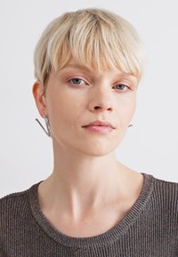 Boucles d'oreilles géométriques en argent avec un design angulaire, mettant en valeur une surface texturée. Portées par un mannequin avec des cheveux blonds clairs et courts, revêtant un pull à côtes.