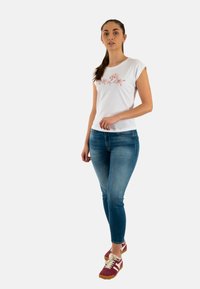 T-shirt bianco a maniche corte con ricami floreali rosa, abbinato a jeans blu attillati e sneaker sportive rosse con accenti bianchi.