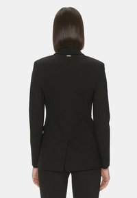 Blazer nero su misura con una silhouette strutturata, spacchetto posteriore singolo e hardware minimalista al colletto. Tessuto di texture liscia.