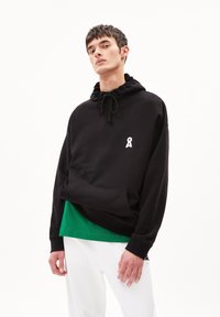 Felpa nera in misto cotone, con tasca a marsupio frontale, cappuccio con cordoncino e logo bianco sul petto. Indossata sopra una maglia verde.