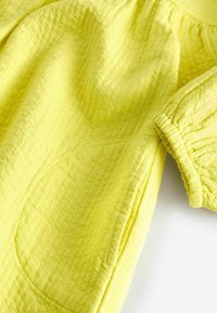 Tissu jaune texturé avec un poignet élastique froncé et une grande poche ovale sur le côté.