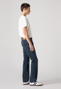 Jeans de perna reta para homem em denim azul escuro com um retalho de couro castanho na cintura, combinados com uma t-shirt branca de mangas curtas e ténis brancos.