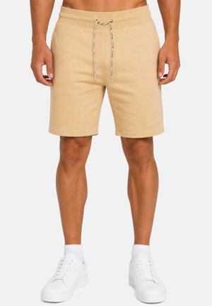 Uomo con pantaloncini beige con coulisse e sneakers bianche, in piedi con le mani rilassate lungo i fianchi davanti a uno sfondo semplice.