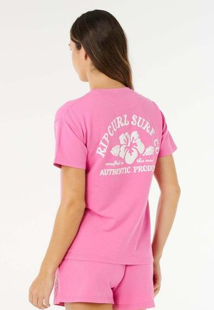 Femme aux longs cheveux portant un t-shirt rose Rip Curl Surf Co. et un short assorti, tournée de dos sur un fond uni.