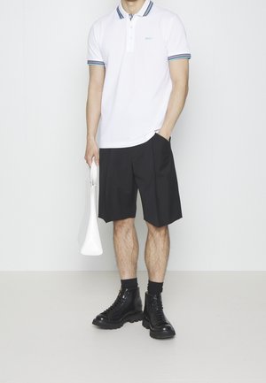 Mann trägt weißes Polohemd, schwarze knielange Shorts, schwarze hohe Stiefel, hält weiße Handtasche in der linken Hand, rechte Hand in der Tasche.