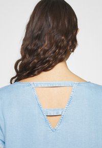 Ljusblå denimtop med en triangulär utskärning i ryggen och horisontella remmar. Slät textur med synliga sömmar längs kanterna.