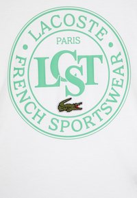 Bílá bavlněná tričko s kruhovým logem v mátové barvě obsahujícím nápisy "LACOSTE," "PARIS," a "FRENCH SPORTSWEAR." Obsahuje vyšívaný detail krokodýla.