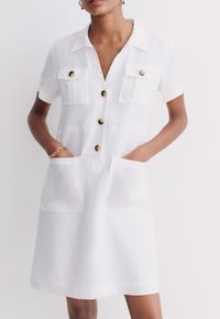 Robe chemise en lin blanc avec manches courtes, deux poches poitrine, deux poches avant et boutons ronds marron. Texture douce, design longueur genoux.