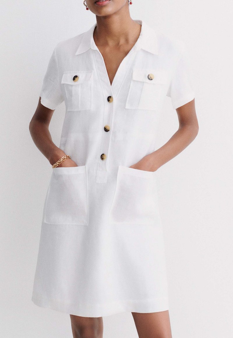 Robe chemise en lin blanc avec manches courtes, deux poches poitrine, deux poches avant et boutons ronds marron. Texture douce, design longueur genoux.