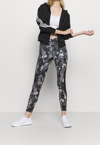 Sudadera negra con cierre y rayas blancas, combinada con leggings ajustados de patrón en negro y gris. La modelo lleva zapatillas deportivas blancas.