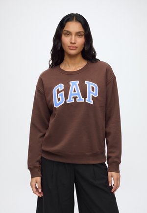 Жена с дълга тъмна коса, носеща кафяв суитшърт с "GAP" в големи сини и бели букви и черен панталон, стояща пред обикновен фон.