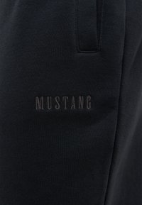 Schwarze Sweatpants aus weichem Stoff, mit dem Wort "MUSTANG" in dunkler Nähte auf der Seite eingeprägt und mit einer glatten, strukturierten Oberfläche.