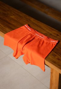 Pomarańczowe bawełniane bokserki rozłożone na drewnianej powierzchni. Na pasku znajduje się biały znak "HAPPY SHORTS", a materiał jest gładki.