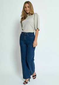 Helles graues Kurzarmoberteil mit gepufften Schultern, kombiniert mit hochgeschnittenen, dunkelblauen weiten Jeans und schwarzen Absatzsandalen.