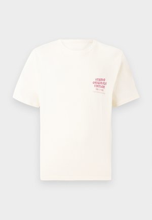 T-shirt blanc à manches courtes avec le texte rouge "Édition Originals du Studio" sur la poitrine gauche, présenté sur un fond gris neutre.