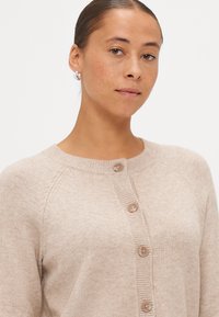 Svetlo bež kardigan iz mehkega pletenega materiala, z okroglim izrezom, petimi gumbi in rebrastimi detajli na manšetam in robu.