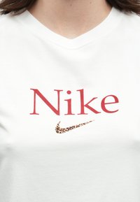 Λεπτομέρεια από ένα λευκό μπλουζάκι με V-λαιμόκοψη, με κόκκινο κείμενο "Nike" και ένα μικρό λογότυπο σήματος με λεοπάρδαλη κάτω από το κείμενο.