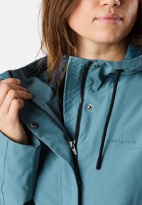 Giacca impermeabile teal con colletto alto, chiusura frontale con zip e lacci regolabili. Presenta ricamo del logo e chiusure a scatto.