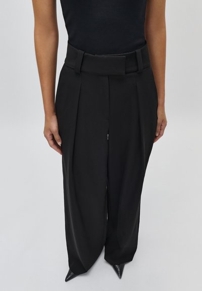 Vrouw met zwarte, hoog getailleerde, wijde broek met riemlussen en puntige zwarte schoenen tegen een effen achtergrond.