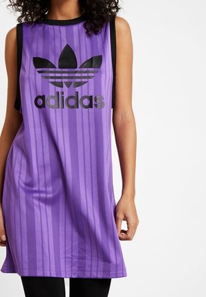 Kobieta w fioletowej, bezrękawnikowej sukience Adidas z czarnym logo i paskami, stoi z jedną ręką luźno opadającą wzdłuż ciała.
