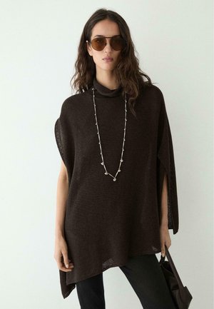 Donna che indossa un poncho lavorato a maglia oversize marrone scuro, lunga collana d'argento, occhiali da sole marroni, tiene una borsa nera contro uno sfondo chiaro.