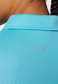 Camiseta polo turquesa con un patrón texturizado de panal y un logo de corazón plateado bordado en la parte posterior del cuello.