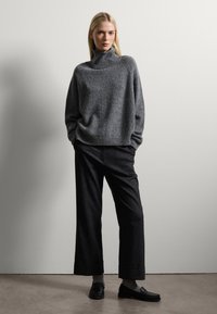 Grijze gebreide trui met hoge kraag, gecombineerd met zwarte wijde broek en zwarte loafers. Soepele textuur met een relaxte pasvorm.