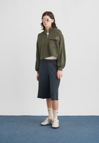 Κοντό φούτερ ελιάς με φερμουάρ και τσέπη, σετ με σκούρο μπλε σορτς. Διαθέτει λευκό λογότυπο, κρεμ κάλτσες και γκρι αθλητικά παπούτσια.