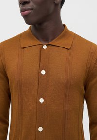 Homme portant une chemise marron en maille boutonnée avec un col pointu, montrant le haut de la poitrine et le cou sur un fond uni.
