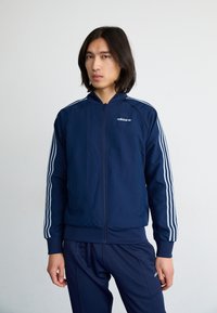 Sötétkék sportdzseki teljes cipzárral, fehér háromsávos mintával a karokon és logóval a mellkason. Puha, sima tapintású.