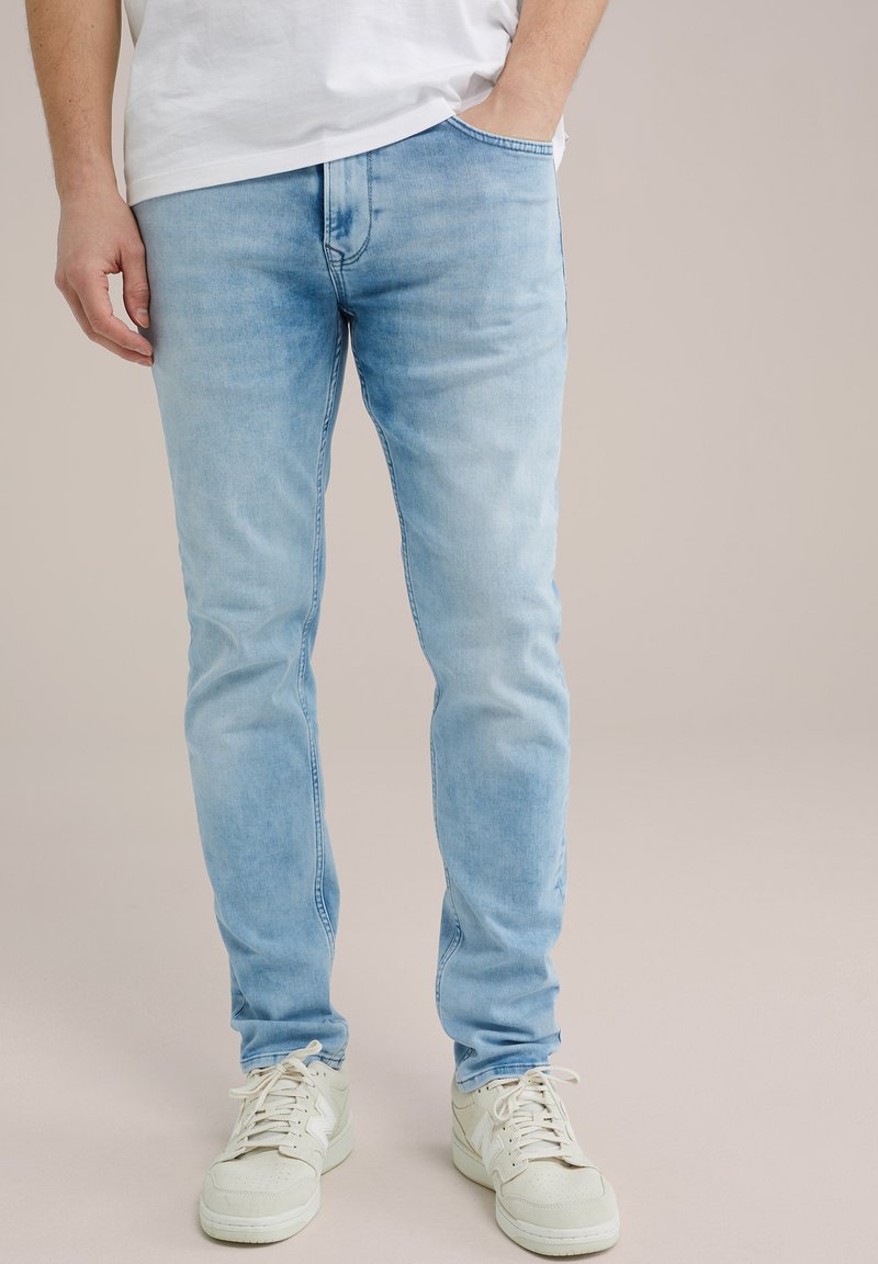 Gaiši zili džinsi ar šauru piegriezumu, ar klasisku taisnu griezumu, niecīgu izbalēšanu un piecu kabatu dizainu, apvienoti ar baltām kedām.