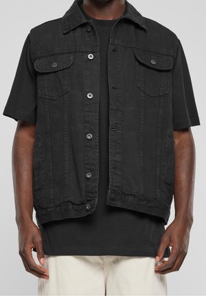 Man draagt een losgeknoopte zwarte mouwloze spijkerjas over een zwart T-shirt met korte mouwen en lichte broek, handen ontspannen langs het lichaam.