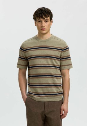 Młody mężczyzna z krótkimi kręconymi włosami, ubrany w beżową, brązową i granatową koszulkę w paski na krótki rękaw oraz brązowe spodnie, stojący na jednolitym tle.