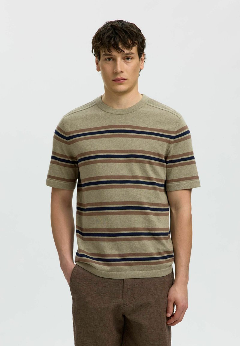 Jeune homme aux cheveux courts et bouclés, portant une chemise à manches courtes rayée beige, marron et bleu marine, et un pantalon marron, debout devant un fond uni.