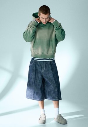 Młody mężczyzna, poprawiający zieloną bluzę z kapturem, w szerokich denimowych krótkich spodenkach, białych skarpetkach i beżowych wsuwanych butach, stojący na jasnym tle.