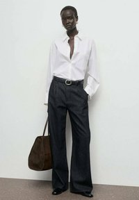 Camisa blanca de botones, pantalones de mezclilla oscuros de pierna ancha, cinturón negro con hebilla circular y bolso de ante marrón. Unos zapatos planos negros completan el look.