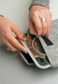 Gray felt pouch with a silver zipper. A hand is opening it to reveal a circular, metallic object inside. Soft texture and minimalist design.