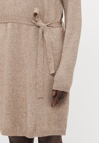 Abito in maglia beige con maniche lunghe, vita cintata e polsini a coste. Il tessuto ha una consistenza morbida, che mostra un sottile motivo melangiato.