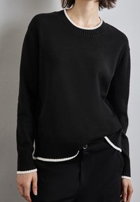 Czarny sweter z prążkowanym okrągłym dekoltem i mankietami, z białym akcentem przy dekolcie, dolnym brzegu i krawędziach rękawów; miękka tekstura.