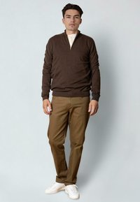 Seis man som står iförd en brun zip-tröja, beige byxor och vita sneakers mot en enkel ljus bakgrund.