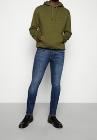 Olivově zelená mikina s kapucí a přední kapsou, se černým logem, ve spojení s modrými slim-fit džínami a černými teniskami. Hladká textilie.