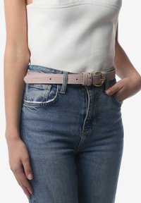 Lyserød læderbælte med krokodillemønster og en gylden cirkulær spænde. Bæltet bæres over højtaljede blå denimjeans. Struktureret overfladedetalje synlig.