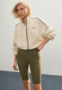 Kremfarvet sweatshirt med lynlås, sorte detaljer og logo, kombineret med olivengrønne leggings der går til låret. Glat tekstur, afslappet pasform.