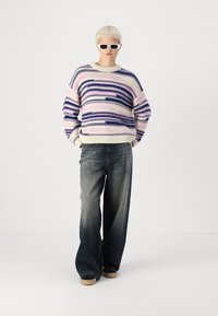 Strikkegenser i pastellrosa, blå og kremfargede striper, kombinert med brede, falmede jeans. Komplettert med hvite solbriller og svarte sko.