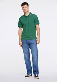 Zielona koszulka polo z białymi wykończeniami, trzy guziki przy kołnierzy, w połączeniu z niebieskimi dżinsami i czarnymi sneakersami. Prosty design, luźny krój.