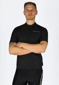 Schwarzes Fahrradtrikot mit kurzen Ärmeln, Reißverschluss vorne, dezentem Branding und einem leichten strukturierten Stoff. Getragen mit enganliegenden schwarzen Shorts.