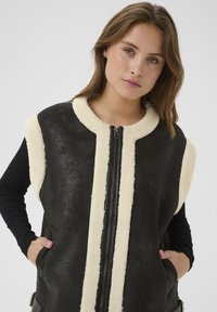 Mlada žena s svijetlosmeškom kosom nosi crni bezrukavnik od ovčje kože s kremastim krznom i crnu dugom rukavicom, s rukama u džepovima.