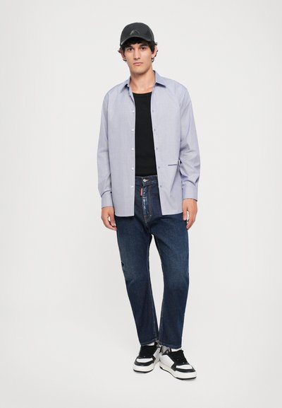 Junger Mann mit schwarzer Mütze, hellblauem Hemd über schwarzem T-Shirt, dunklen Jeans und schwarz-weißen Sneakers, der vor einem weißen Hintergrund steht.