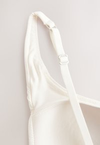 Nahaufnahme eines weißen verstellbaren BH-Trägerbands und der Körbchenkante mit Nahtdetails auf hellem Hintergrund.